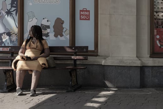 Windsor, UK - Apr 03, 2025 - African American women or Black women Wearing a yellow dress sitting on on a wooden bench in front of Miniso shop and holding a phone in hand. use it as your Wallpaper, Poster and Space for text, Selective focus.