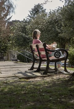 Windsor, UK - Apr 03, 2025 - Side view of A middle-aged woman wear glasses and a cream-colored shoulder bag sitting relax on a wooden bench in the park of church. use it as your Wallpaper, Poster and Space for text, Selective focus.