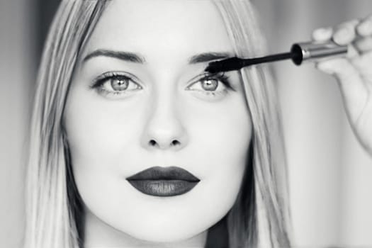 Close-up of a beautiful blonde woman applying mascara, with dark lipstick, bright eyes, and long straight hair. Black and white portrait for beauty, makeup, fashion, or cosmetic themes