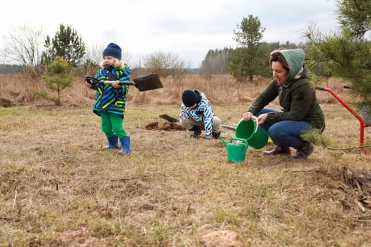 A woman assists two children as they plant young trees in an open grassy area. The children are using shovels and watering cans while dressed warmly for the cool weather.