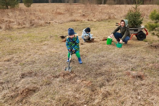 On a sunny day, two children and an adult work together in a grassy field. They are digging holes and planting young trees while enjoying the fresh air and nature.