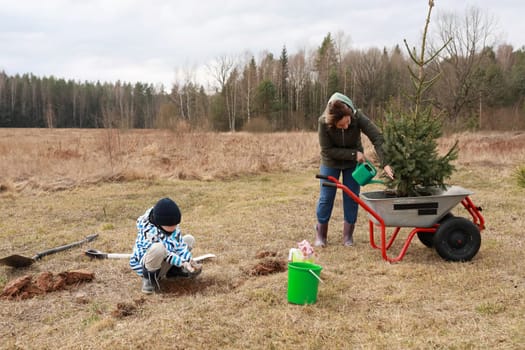 Two individuals engage in planting a tree in a grassy area. One child digs the ground while an adult waters the tree nearby. The day is overcast, hinting at early spring.
