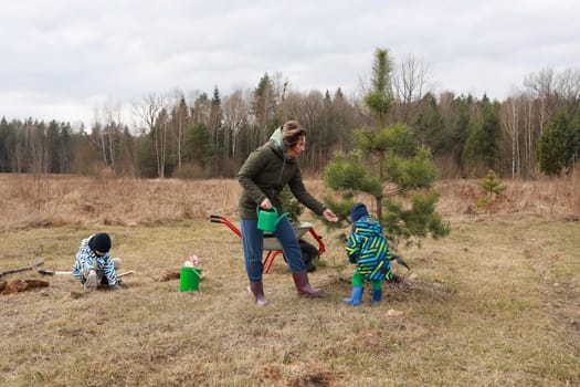 A woman helps two children care for young trees in a field near a forest. One child plants, while the other waters a tree, showcasing teamwork in nature.