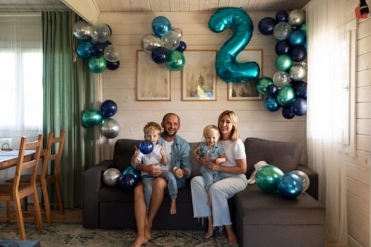 A family enjoys a birthday party at home, celebrating a child's second birthday with colorful balloons and a playful atmosphere. The living room is warmly decorated.