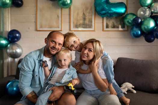 A family enjoys a joyful moment together in a cozy living room filled with blue and green balloons. Two children play with toy cars while smiling closely with their parents.