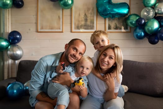A family of four gathers on a couch, surrounded by colorful balloons. Two children play with toy cars while parents smile, enjoying a joyous moment at home in the afternoon.