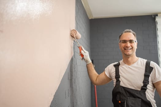 A man is standing in front of a bare wall, actively painting it with a paint roller. He is focused on his task, applying a fresh coat of paint smoothly and evenly.