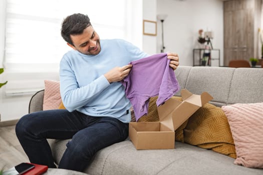 Brown-haired man sits on the couch in the living room and happily opens a cardboard package and looks at a new T-shirt.