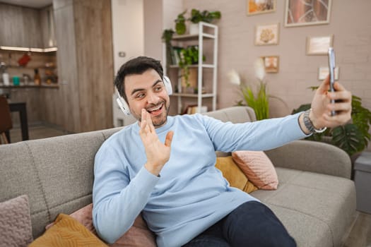 Brown-haired man with white headphones on his head sits on the couch in the living room and makes a video call on a mobile phone.....
