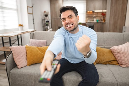 A man is seated on a couch, holding a remote control in his hand. He appears to be focused on the TV screen. The room is dimly lit, with a cozy atmosphere.