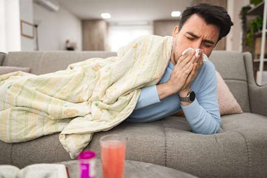 Man is seen laying on a couch, covering his face with a tissue, as he blows his nose due to sickness or allergies. His eyes may appear watery or tired.