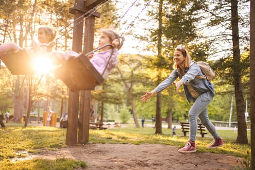 Mother is rocking her twin daughters on a swing in the park.