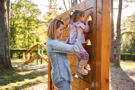 Smiling mom helps her cute daughter climb the playground.