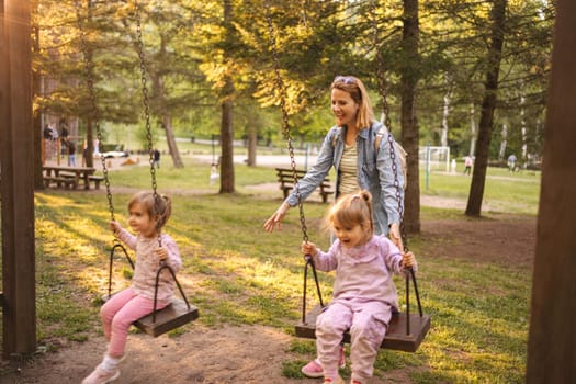 Mother is rocking her twin daughters on a swing in the park.