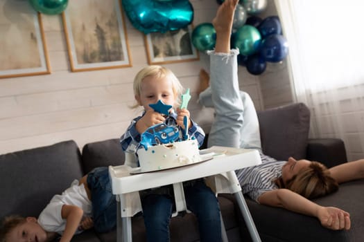 A toddler engaged in eating cake sits in a high chair while a sibling and parent relax on the couch. Colorful balloons decorate the cozy living room, creating a festive atmosphere.