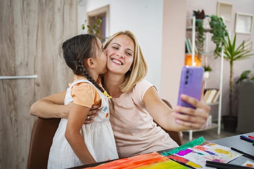 Daughter kisses her mother as they happily capture a selfie together in their kitchen.