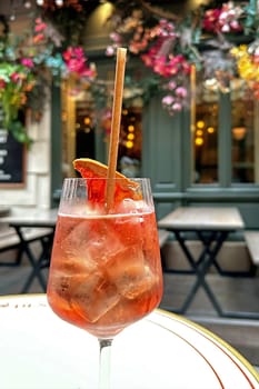 A vibrant Aperol Spritz cocktail, glistening with ice and garnished with an orange slice, stands elegantly in a stemmed glass on a Parisian cafe table. The warm, inviting ambiance is captured with soft focus, showcasing the cafe's exterior adorned with colorful flowers and rustic tables and chairs. This image evokes the feeling of a leisurely afternoon in Paris, perfect for lifestyle, travel, and food-related content.