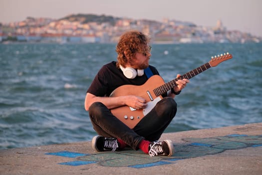 Hipster street musician in black playing electric guitar in the street on sunset on embankment with Lisbon in background. Portugal