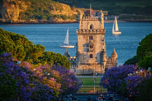 Scenic view of Belem Tower in Lisbon, Portugal, seen over a street with blooming purple jacaranda flower trees street with tourist sailboats on the Tagus River on sunset. Portugal