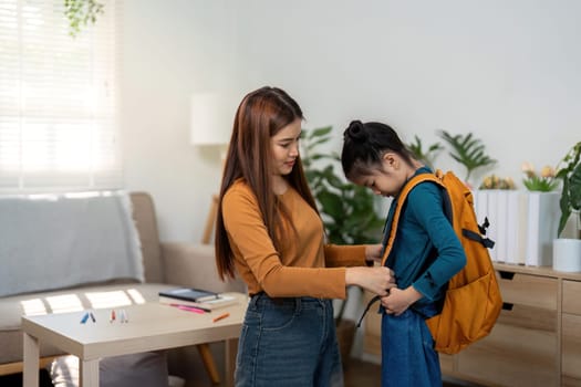 The mother assists her daughter in putting on her backpack, preparing her for a day at school, showcasing nurturing parenting.