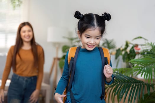 A cheerful young girl wears a bright backpack as she walks towards the door, ready for school. Her smiling mother observes, highlighting the warmth of family support.