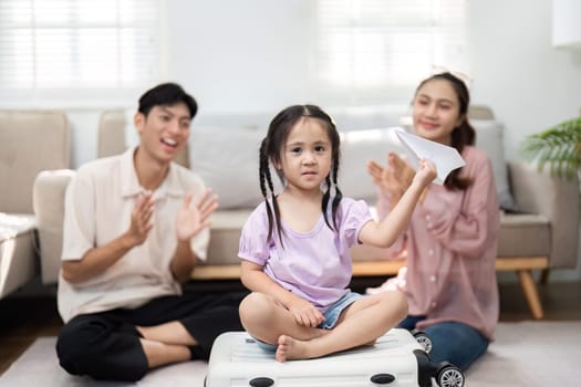In a cozy living room, a young girl sits on a suitcase, showcasing her handmade paper airplane to her happy parents. The scene highlights family connection and playful encouragement.