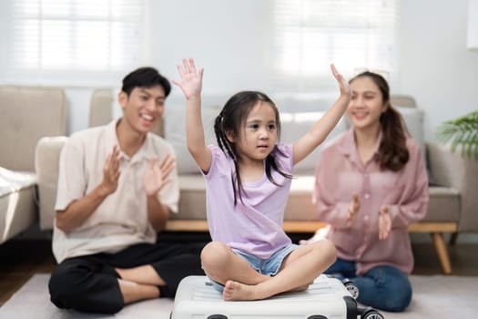 In a lighthearted moment, a girl raises her hands in excitement while sitting on a suitcase, as her joyful parents celebrate the fun of preparing for their family journey.