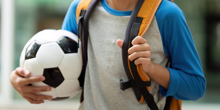 A young boy with a soccer ball is accompanied by his mother as they walk towards school, sharing smiles and excitement about the day ahead.