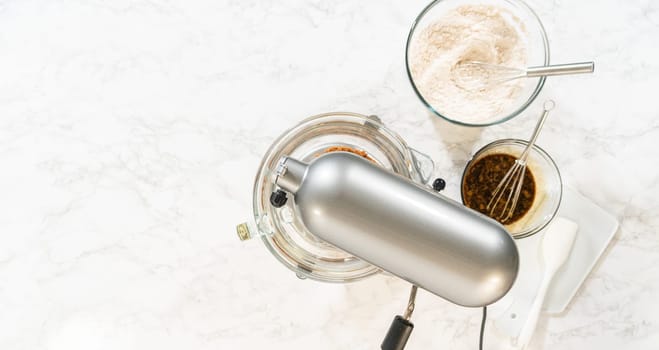 Overhead view of a baking setup for Classic Gingerbread Cookie Dough, with a stand mixer surrounded by essential ingredients. Ingredients include brown sugar, butter, flour mixture, and dark molasses in individual glass bowls, showcasing the classic components needed for this festive dough recipe.
