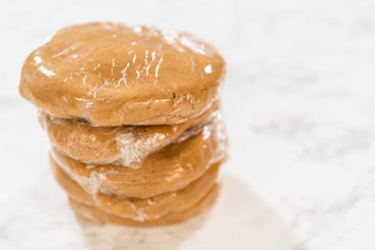 Stacked view of multiple discs of Amber Gingerbread Cookie Dough, each individually wrapped in plastic wrap, ready to be stored or chilled. The layers are neatly stacked, showing careful preparation for later baking.