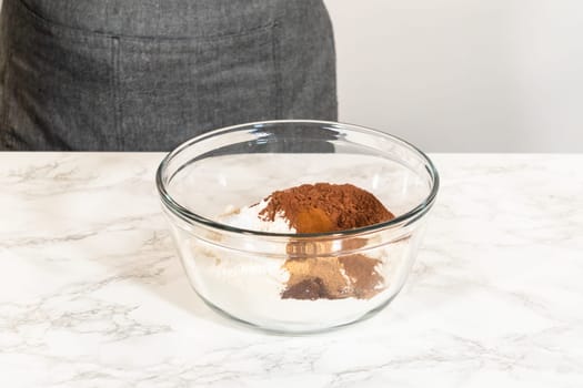 A bowl containing dry ingredients for Chocolate Gingerbread Cookie Dough, including cocoa powder, spices, and flour, ready to be mixed on a marble countertop.