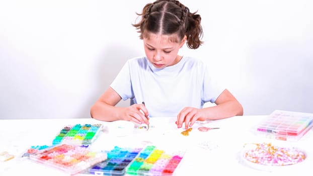 Delicate fingers of a young girl navigate through a treasure trove of bright, multicolored beads, each compartment revealing a new hue to choose from. She's immersed in the joyful task of stringing together a handmade bracelet, with golden beads and pearls lying nearby to add a touch of sparkle to her creation.