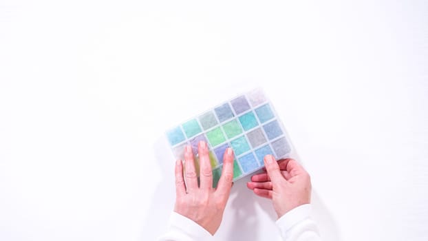 Flat lay. Woman hands gracefully poised over a collection of beads, sorted by color in transparent organizers. The array of beads spans a vibrant spectrum, from deep purples to bright oranges, meticulously arranged for easy selection as she embarks on creating a custom piece of jewelry.
