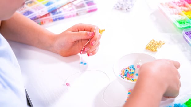 Delicate fingers of a young girl navigate through a treasure trove of bright, multicolored beads, each compartment revealing a new hue to choose from. She's immersed in the joyful task of stringing together a handmade bracelet, with golden beads and pearls lying nearby to add a touch of sparkle to her creation.