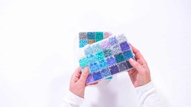 Flat lay. Woman hands gracefully poised over a collection of beads, sorted by color in transparent organizers. The array of beads spans a vibrant spectrum, from deep purples to bright oranges, meticulously arranged for easy selection as she embarks on creating a custom piece of jewelry.
