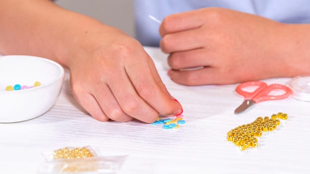Delicate fingers of a young girl navigate through a treasure trove of bright, multicolored beads, each compartment revealing a new hue to choose from. She's immersed in the joyful task of stringing together a handmade bracelet, with golden beads and pearls lying nearby to add a touch of sparkle to her creation.