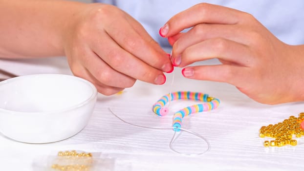 Delicate fingers of a young girl navigate through a treasure trove of bright, multicolored beads, each compartment revealing a new hue to choose from. She's immersed in the joyful task of stringing together a handmade bracelet, with golden beads and pearls lying nearby to add a touch of sparkle to her creation.
