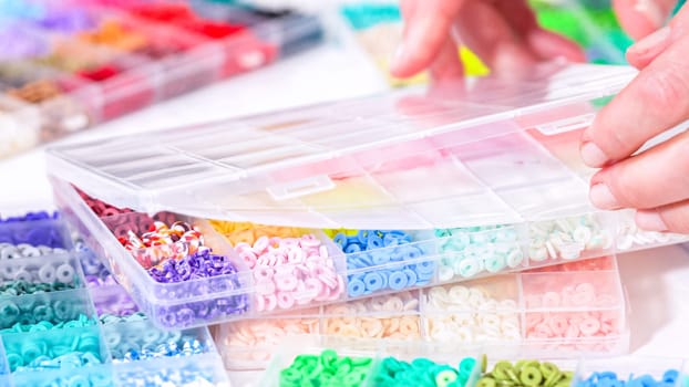 Woman hands gracefully poised over a collection of beads, sorted by color in transparent organizers. The array of beads spans a vibrant spectrum, from deep purples to bright oranges, meticulously arranged for easy selection as she embarks on creating a custom piece of jewelry.