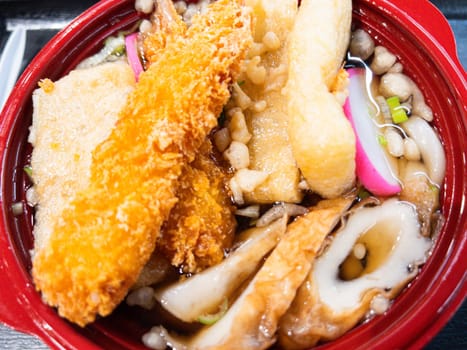 A bowl of Japanese udon noodle soup topped with crispy tempura, fish cakes, fried tofu, and green onions. The dish is served in a red bowl with a rich broth and crunchy tempura flakes, creating a flavorful and comforting meal.