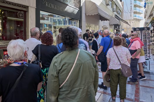 Crete, Greece - April 7, 2025: Lively urban atmosphere featuring tourists enjoying vibrant shopping streets in Crete. They are casually dressed, surrounded by shops, a display stand, and welcoming storefronts