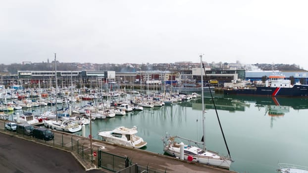 Boulogne. France 03.28.2025 Overcast view of a marina filled with sailboats and yachts, bordered by fencing and industrial buildings in the background.