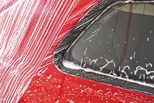 Red car washed in self serve carwash, detail on white soap foam flowing over side window and red body plates