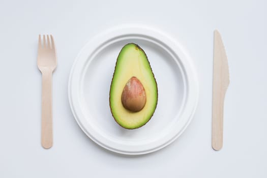 Minimalist composition with avocado halve on white plate and wooden cutlery.