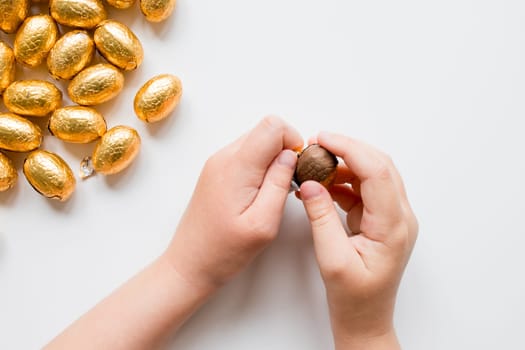 Child opening chocolate egg with gold foil candy on white background.