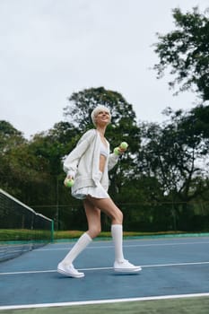 A joyful young woman in a sporty outfit plays tennis outdoors on a sunny day, smiling and holding tennis balls, with a vibrant green background, perfect for sports and active lifestyle themes