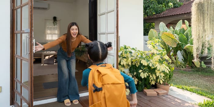 A cheerful mother opens her arms wide to greet her daughter, who is returning home from school, highlighting the joy of family togetherness.