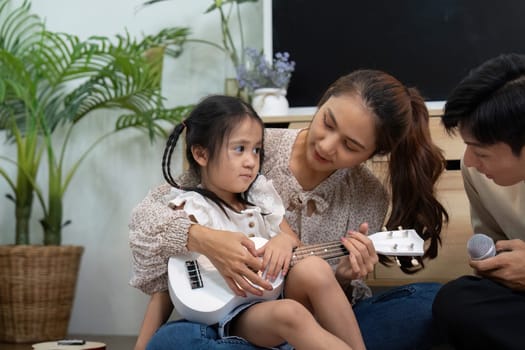A close-knit family enjoys a musical moment, where a mother patiently guides her daughter on the ukulele while sharing smiles and laughter, enhancing their bond.