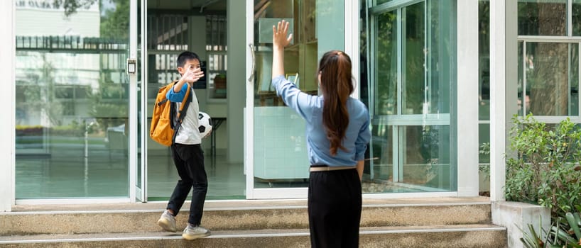 The young boy waves back to his mother while entering school, showcasing the bond and support between them, and the excitement of school life.