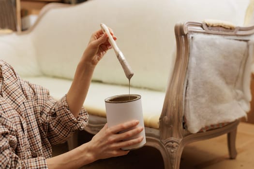 Close-up of woman's hands holding a paintbrush dripping with beige paint, restoring vintage furniture at home.