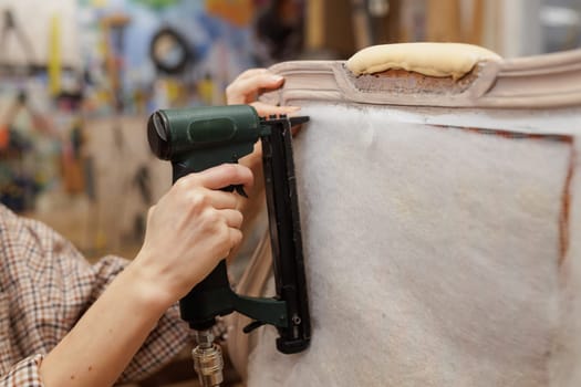 Close-up of hands using a pneumatic stapler to attach padding on a vintage armchair during upholstery work.